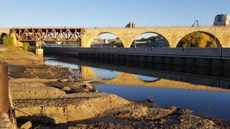 View of rocky Mississippi River river bottom after water level was lowered with Stone Arch Bridge and railroad bridge in the background